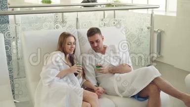 穿着浴袍的年轻情侣们在温泉沙龙的椅子上放松，喝鸡尾酒，聊天和接吻。 健康