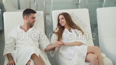 年轻夫妇中<strong>有</strong>魅力的女人和穿着浴袍的坐在日间水疗中心的放松室里，手牵手，