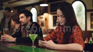 年轻的女学生正在使用智能手机，坐在高档酒吧和鸡尾酒。 她摸着屏幕微笑着。 现代