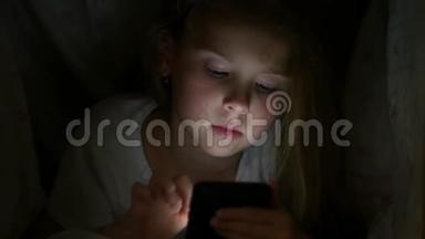 女孩晚上在毯子下看手机。 夜间智能手机游戏