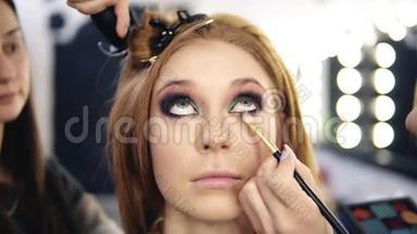 一个金发美女模特坐着抬头看着化妆师整理她烟熏的眼睛的镜头
