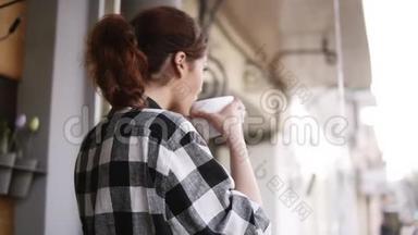 一个穿着格子衬衫的可爱而高兴的女孩站在窗户对面，喝着一杯白色的大杯子，向外望去