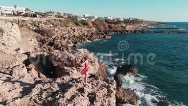 穿着红色衣服的女孩迎风招展，双手分开在岩石悬崖上，海浪拍打着海岸，溅起浪花