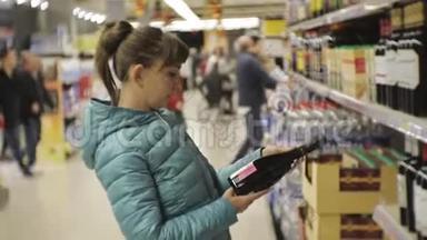 超市里的女人。穿着蓝色夹克的年轻白种女人在阅读深色瓶子上的标签，选择<strong>起泡酒</strong>。