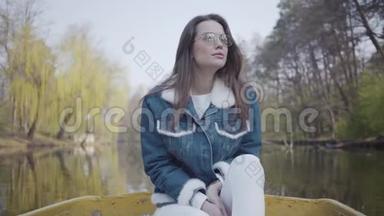 一幅美丽的年轻女子戴着<strong>太阳</strong>镜和一件牛仔夹克漂浮在湖面或<strong>河流</strong>上的船上的肖像。 美丽美丽