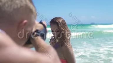 男子摄影师拍摄年轻女子比基尼在海边摆姿势。 年轻的摄影师