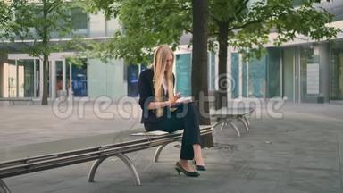 女人在长凳上用平板电脑。 正式的女商人坐在办公室露台的长凳上，休闲地浏览平板电脑。