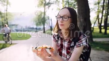 快乐的学生女孩吃了一片披萨，微笑着坐在街上的公园里吃东西。