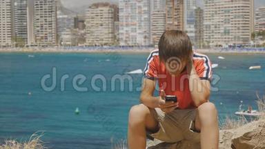 白人少年坐在石头上看着海上摩天大楼天际线背景上的智能手机屏幕。