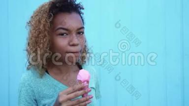 年轻的黑人女孩吃着粉红色的冰淇淋，微笑着看着蓝墙背景