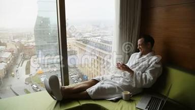 年轻英俊的商人穿着<strong>白色浴袍</strong>坐在床上，手提电脑和手机在现代的肖像