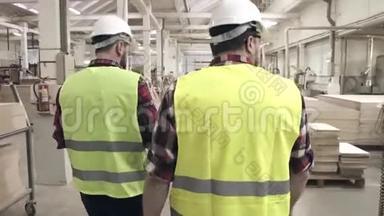 在一家家具厂，两名工人穿着绿色背心和头盔在机器间交谈