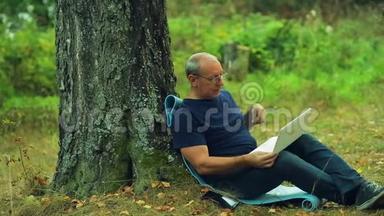 一个戴眼镜的人坐在公园的一棵树下，用笔记本电脑工作。