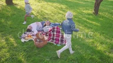 父母躺在草地上，他们的两个女儿在公园里玩得很开心，也有跑步的背景。 家庭度<strong>假景观</strong>。