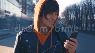 英俊的年轻人在阳光城市街道城市背景。 穿红色连帽衫的人用智能手机，日落背景。