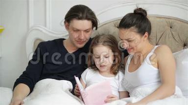 漂亮的小女孩正试图和她的父母躺在床上看书。 美丽的孩子正在用手指朗读