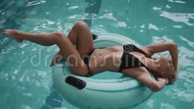 迷人的年轻金发瘦女人穿着<strong>黑色比基尼</strong>躺在游泳池里的充气橡胶圈上。 4k.