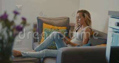 高质量的4k12位镜头，美丽的女孩使用平板电脑坐在家里的沙发上。 女人上网，社交
