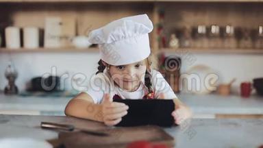 可爱的小女孩在早餐时使用智能手机坐在厨房里。 她穿着围裙，看着
