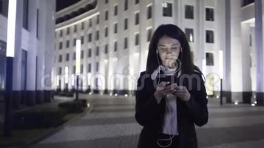 年轻的女商人带着智能手机和耳机走在城市的一条<strong>夜晚街道</strong>上