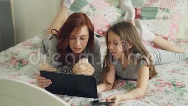 美丽的年轻妈妈和她可爱的女儿穿着睡衣躺在床上笑着看着数码平板电脑