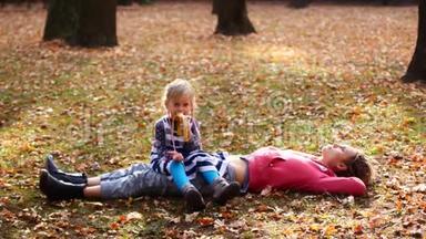 美丽的妈妈和可爱的孩子躺在秋天公园的草地上。 女儿坐在妈妈身上吃香蕉，妈妈望着天空