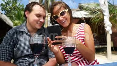 年轻快乐的夫妇喝葡萄<strong>酒</strong>，使用智能手机在户外海滩咖啡馆休息。 慢动作。 1920x1080