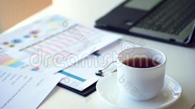 在一个白色的桌面上，有一杯白色的茶，旁边有一台笔记本电脑，并展开商业图表
