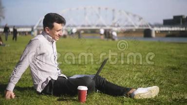 一个人坐在草地上的公园里。 一个自由职业者做得很好，休息，微笑。 夏天。 有笔记本电脑的人。
