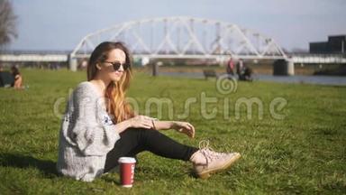 年轻的时髦女人坐在绿草上喝热咖啡，早上享受新鲜空气。 享受外卖咖啡的女人