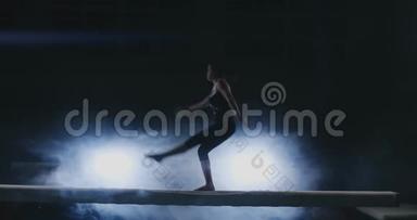这个女孩在一个背光的日志上表演一个技巧，在运动体操服装中表演慢动作。 烟和蓝色。 跳跃和跳跃