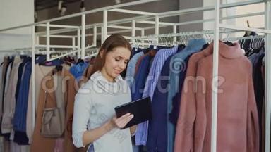 年轻的女商人在她的服装店检查商品时使用平板电脑。 助理带来了服装和