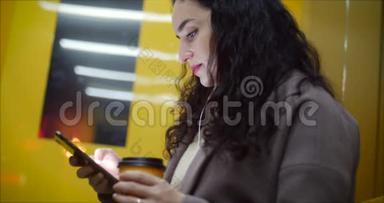 漂亮的女人坐在公共汽车站，用智能手机喝咖啡，在后台有夜城。