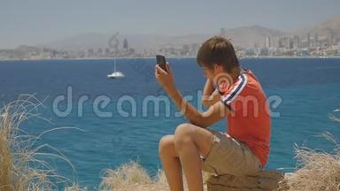 15岁的小男孩穿着红色t恤和短裤，在海滨城市天际线背景下制作智能手机视频电话。
