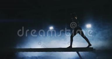 这个女孩在一个背光的日志上表演一个技巧，在运动体操服装中表演慢动作。 烟和蓝色。 跳跃和跳跃