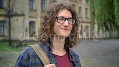 年轻英俊的快乐学生戴着眼镜和背包，站在大学校园附近的街道上，微笑着