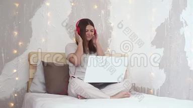 穿着睡衣的漂亮女孩坐在床上用手机听耳机听音乐。 高清高清