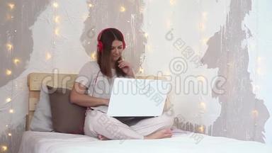 快乐美丽的女孩穿着睡衣，用手机电脑坐在床上听耳机上的音乐。 高清高清