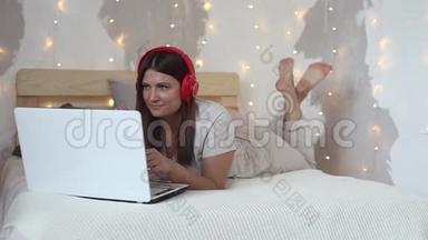 美丽快乐的女孩躺在一张大床上，用一台白色的移动电脑，用耳机听音乐