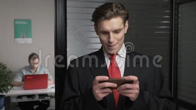 年轻成功的商人穿西装打字，使用智能手机，在智能手机上玩。 男人在电脑上工作