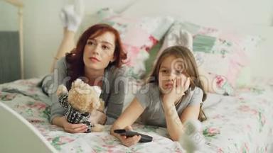 快乐的母亲和可爱的女儿在电视上看卡通电影，一边用遥控器和微笑躺在床上。