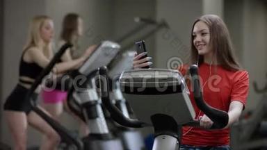 可爱的红衬衫女孩在运动自行车上大力工作，并与她的手机在新的健身房对抗