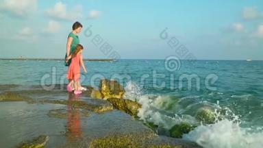 妈妈和女儿站在海<strong>水中</strong>潮湿的岩石上，周围有飞溅的<strong>波浪</strong>，玩得很开心。 慢动作