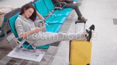 疲惫的女人正坐在机场候机大厅的座位上休息，双腿搭在黄色的手提箱上，看着磁带新闻