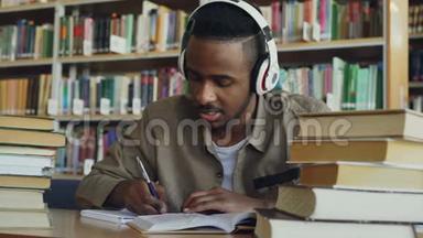 英俊的非裔<strong>美国</strong>男学生戴着耳机，坐在宽敞的<strong>图书馆</strong>里的桌子旁听音乐