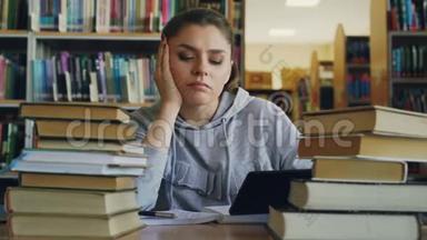 美丽的白种人少女坐在大学图书馆的桌子上，手里拿着数字平板电脑。 她很累，而且