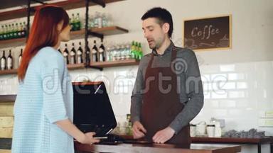 迷人的年轻女子正在和友好的男收银员交谈，她们在咖啡店用手机支付外卖咖啡的费用