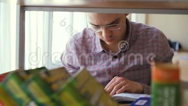 年轻英俊的白种人男学生戴着眼镜站在书架前拿着翻页的书