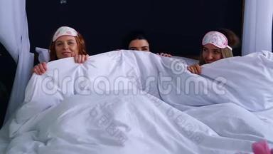 三个女孩在一条毯子下面凌乱地穿着睡衣下床，微笑着看着对方。