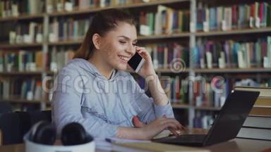 美丽微笑的白种人女学生坐在<strong>大图</strong>书馆的桌子旁，在智能手机上聊天，看着笔记本电脑
